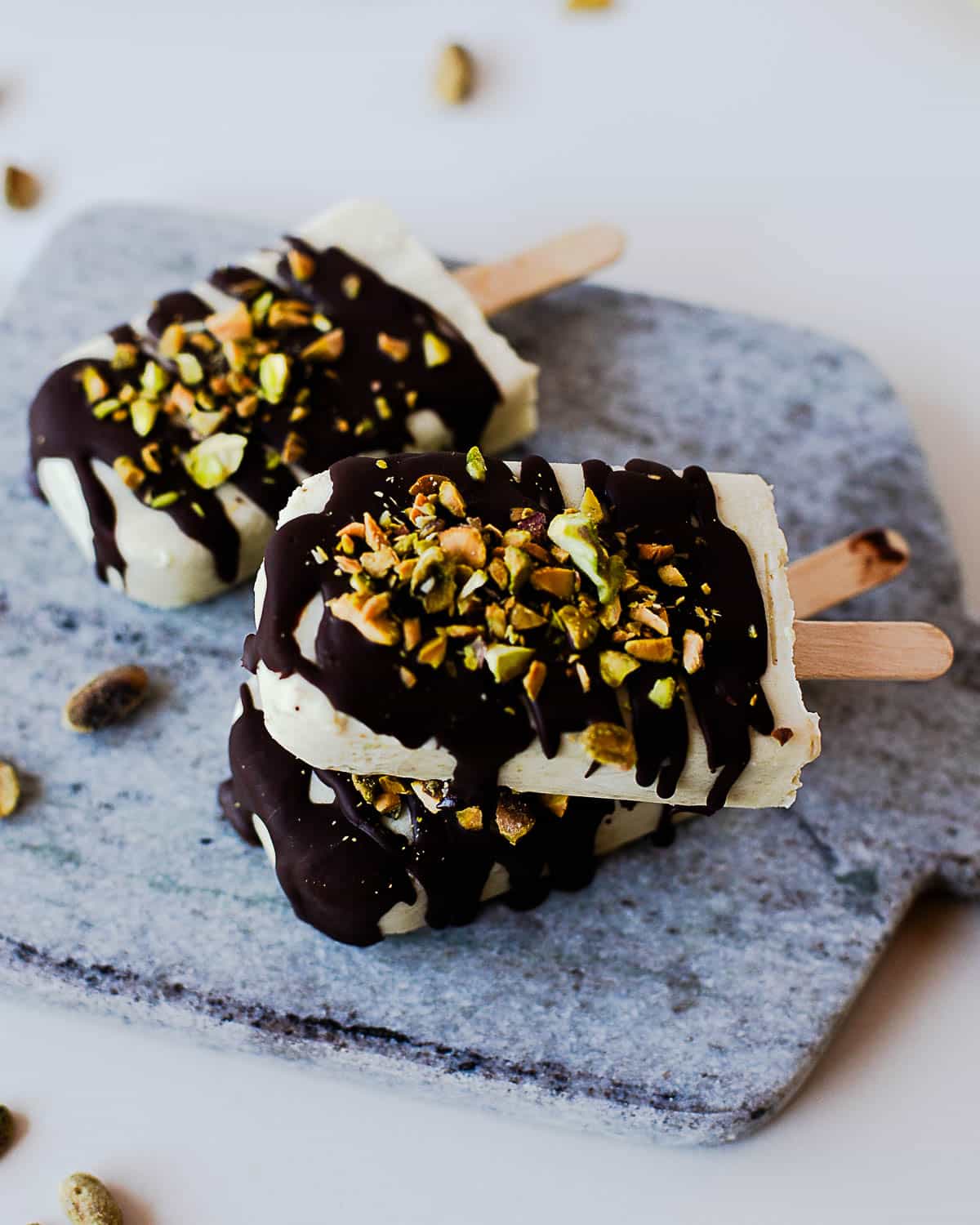 Chocolate drizzled pistachio popsicles stacked on a stone serving board with chopped pistachios.