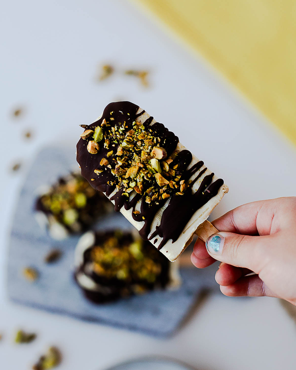 Hand holding a homemade pistachio popsicle topped with dark chocolate and chopped pistachios.