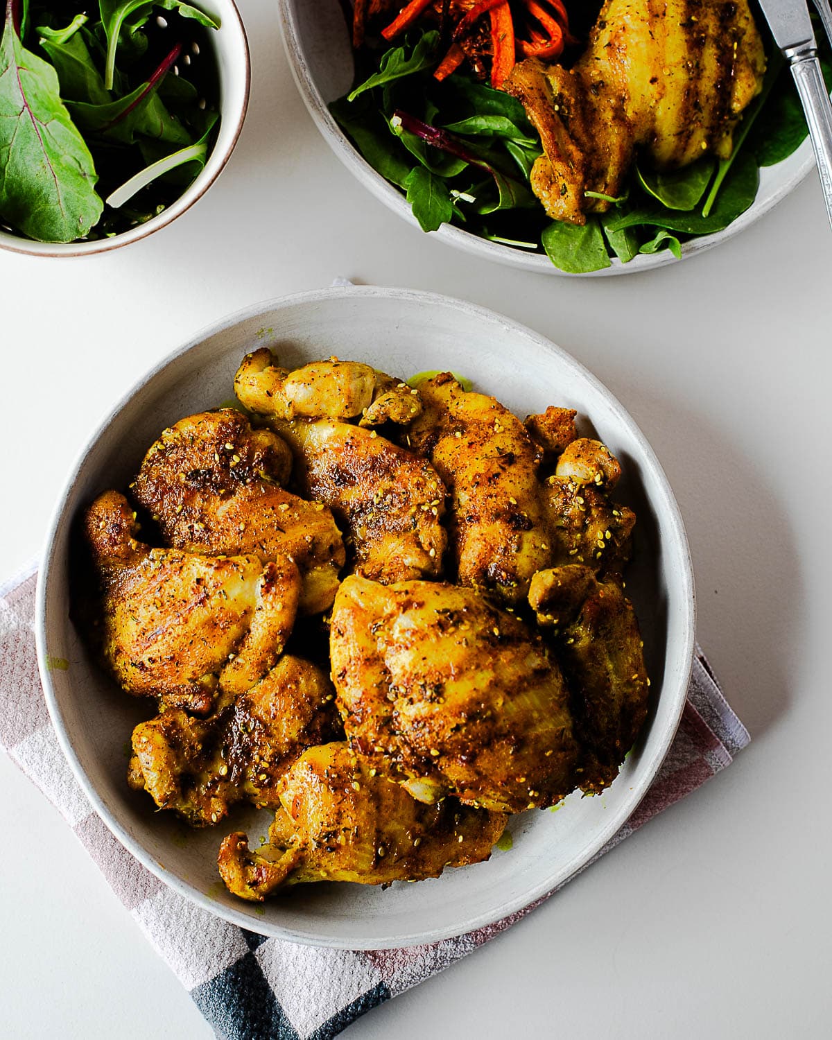 Bowl of cooked za’atar chicken thighs with a simple green salad in the background.
