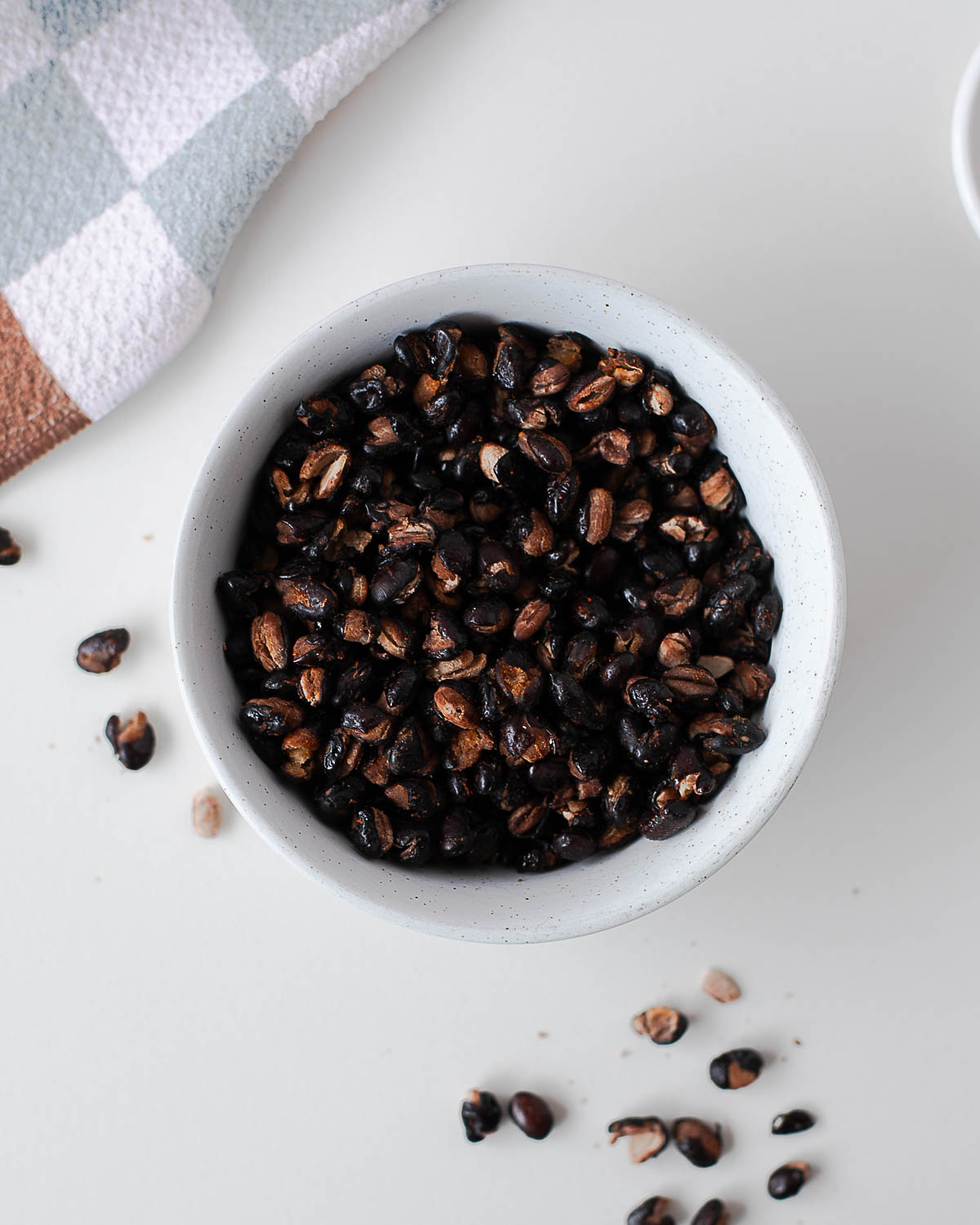 Overhead view of crispy roasted black beans in a small speckled bowl next to a checkered kitchen towel.