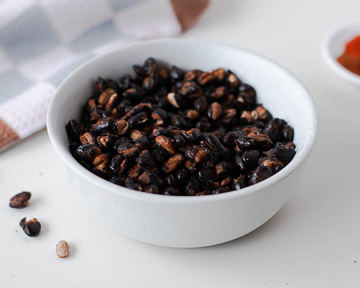 Bowl of crispy roasted black beans on a white countertop with a dish of chili seasoning blurred in the background.