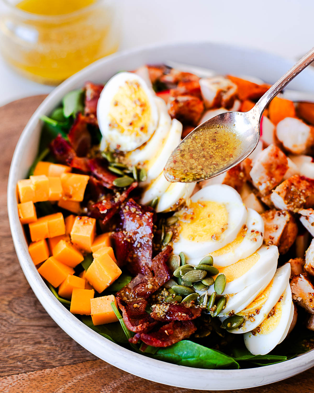 Close-up of a spoon drizzling maple-dijon dressing over sliced eggs and pumpkin seeds on a harvest cobb salad