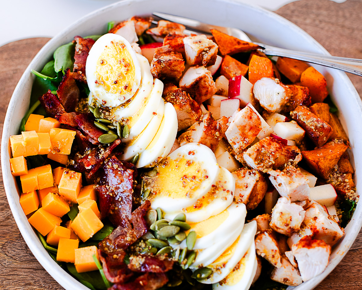 Overhead close-up of harvest cobb salad in a white bowl with rows of cheddar, bacon, sliced eggs, pumpkin seeds, chicken, apple, and roasted sweet potato over spinach