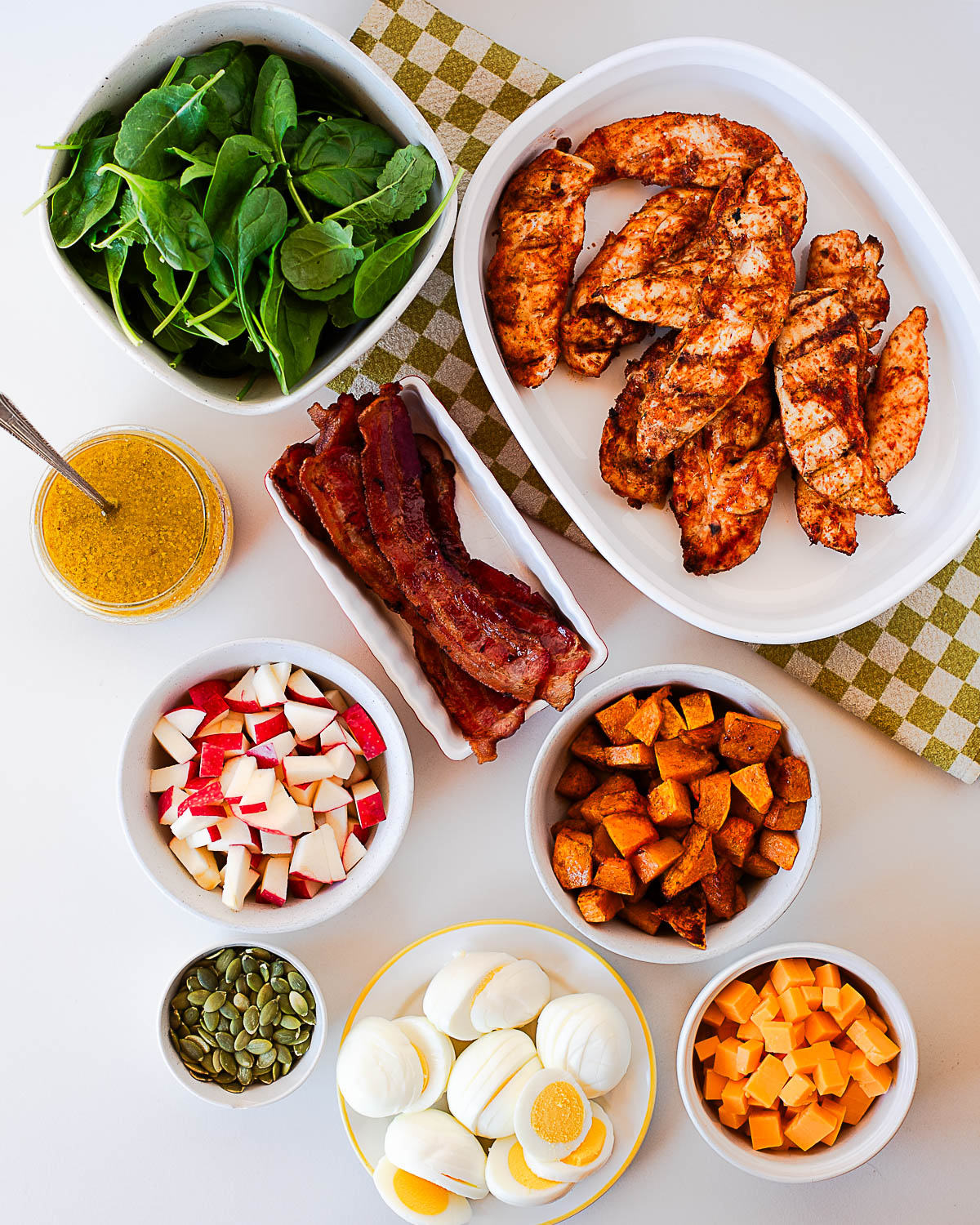 Overhead flat lay of harvest cobb salad ingredients including spinach, seasoned chicken tenders, crispy bacon, roasted sweet potatoes, diced apple, hard-boiled eggs, pumpkin seeds, cubed cheddar, and a jar of maple-dijon dressing