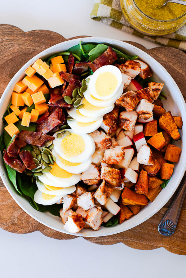 Overhead shot of harvest cobb salad on a wood serving board with a jar of maple-dijon dressing and checkered napkin in the background