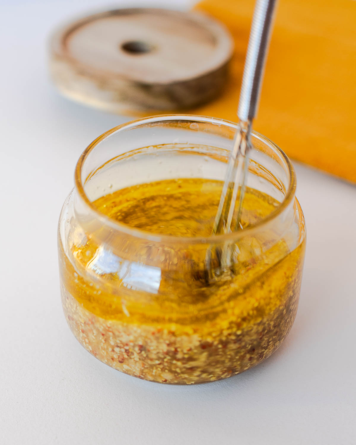 Close-up of a whisk stirring maple mustard dressing in a glass jar, with a wood coaster and mustard-yellow napkin in the background