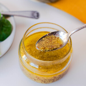 Close-up of a spoon lifting maple-dijon dressing from a glass jar, with spinach and a wood board in the background