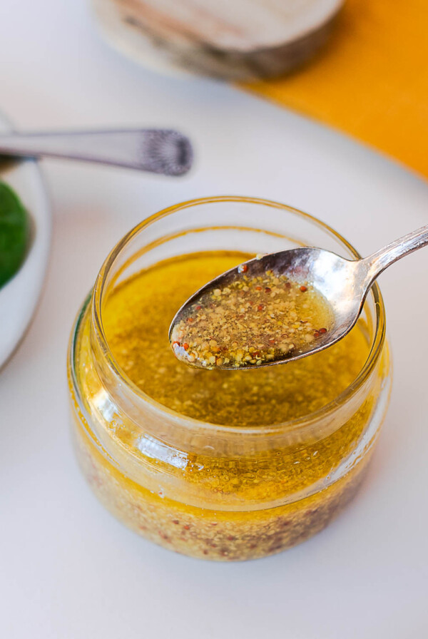 Close-up of a spoon lifting maple-dijon dressing from a glass jar, with spinach and a wood board in the background