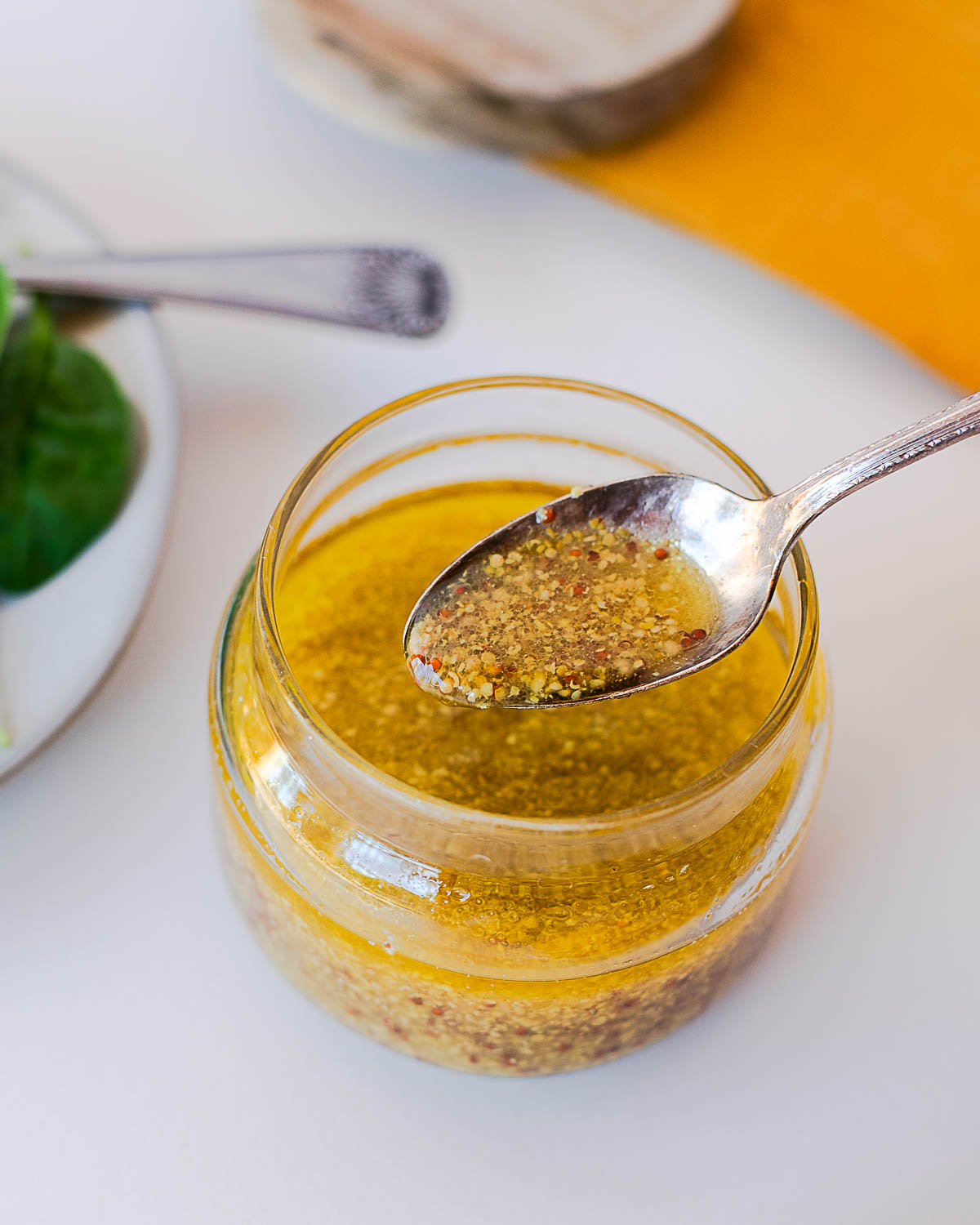 Close-up of a spoon lifting maple-dijon dressing from a glass jar, with spinach and a wood board in the background