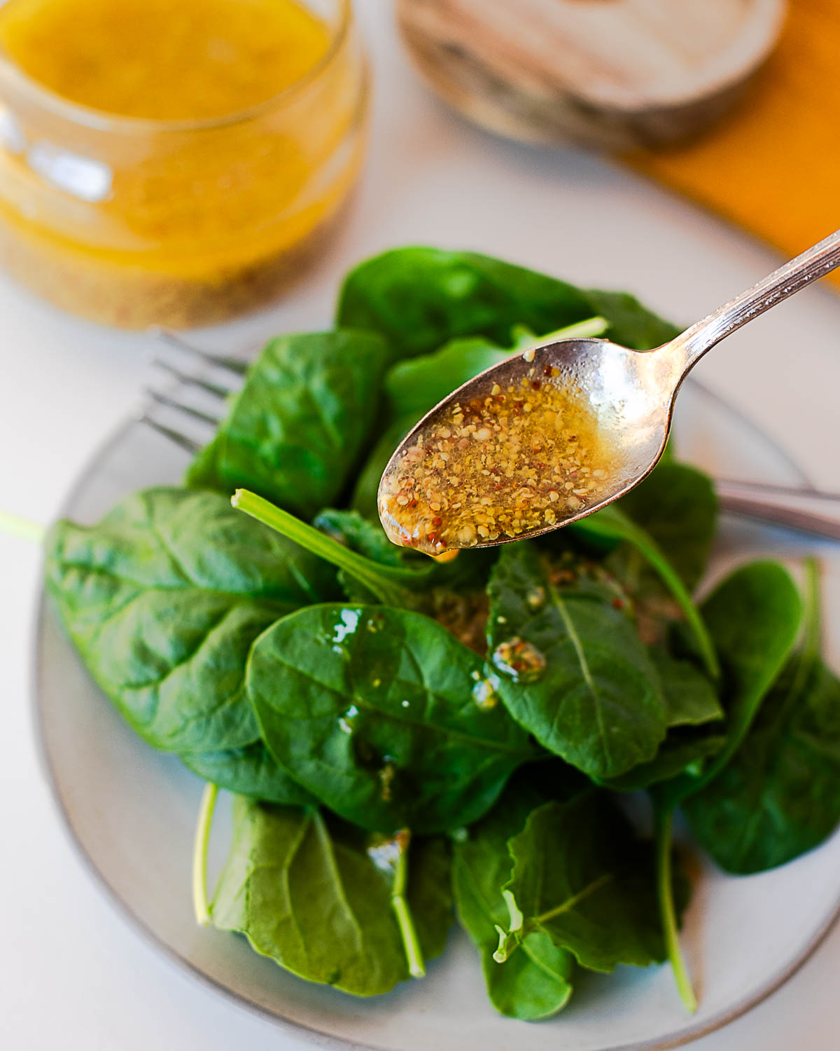Close-up of a spoon drizzling maple mustard dressing over a plate of fresh spinach leaves