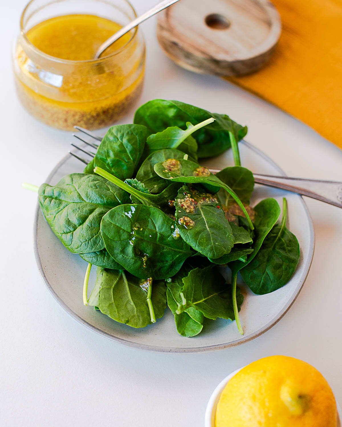 Overhead shot of a plate of spinach drizzled with maple mustard dressing, next to the jar of dressing and a lemon