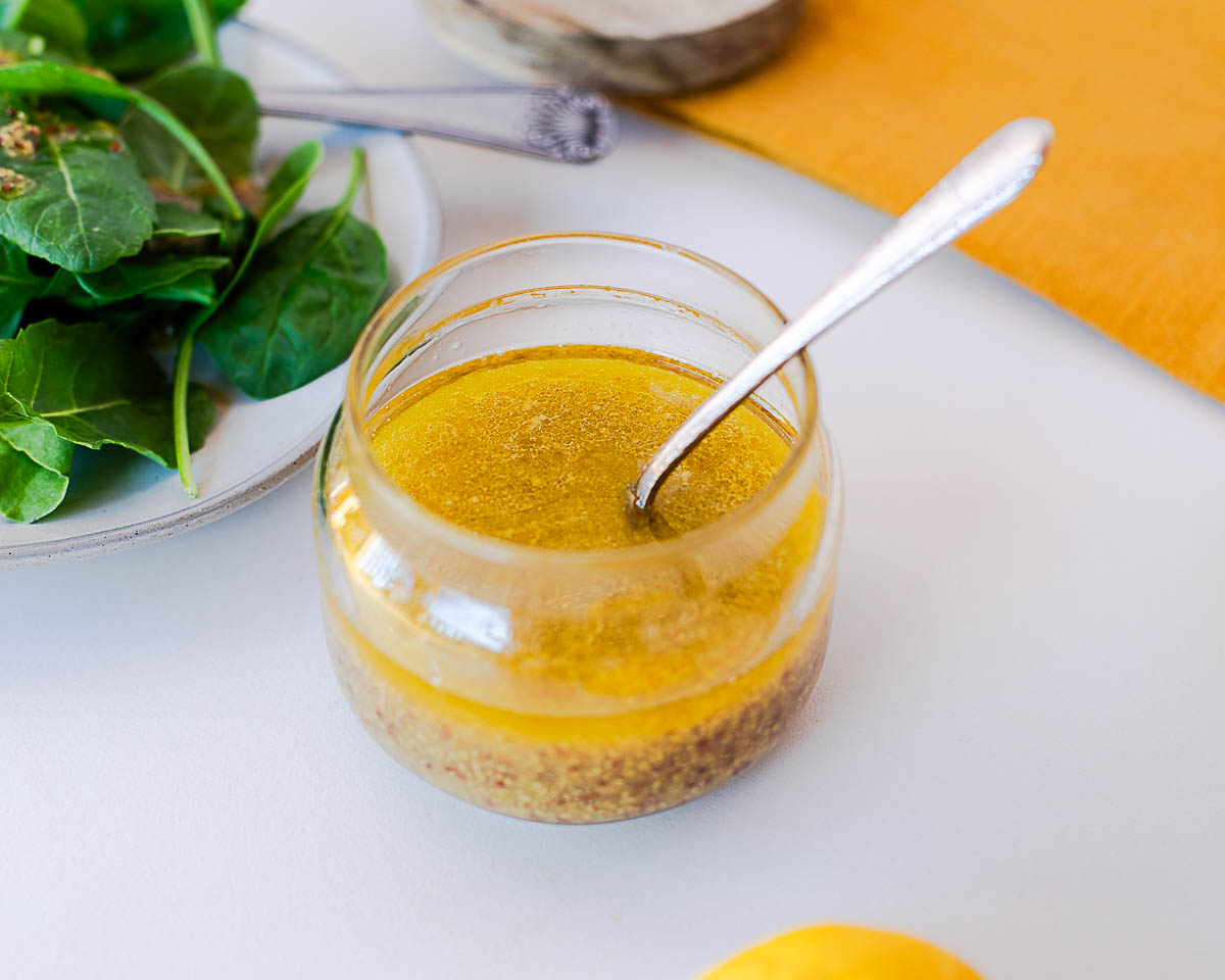 Overhead shot of maple mustard dressing in a glass jar with a spoon resting inside, spinach and a lemon nearby