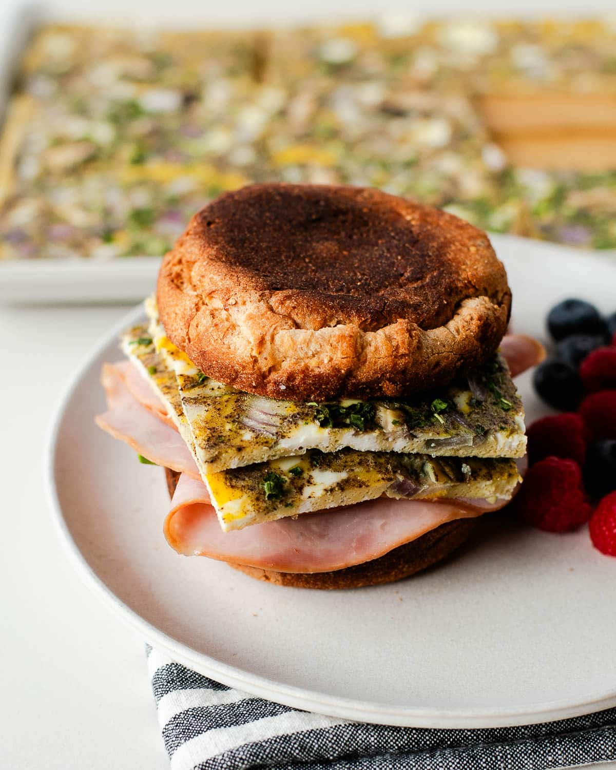 Side view of a breakfast egg sandwich with layered egg bake and ham, berries in the background.