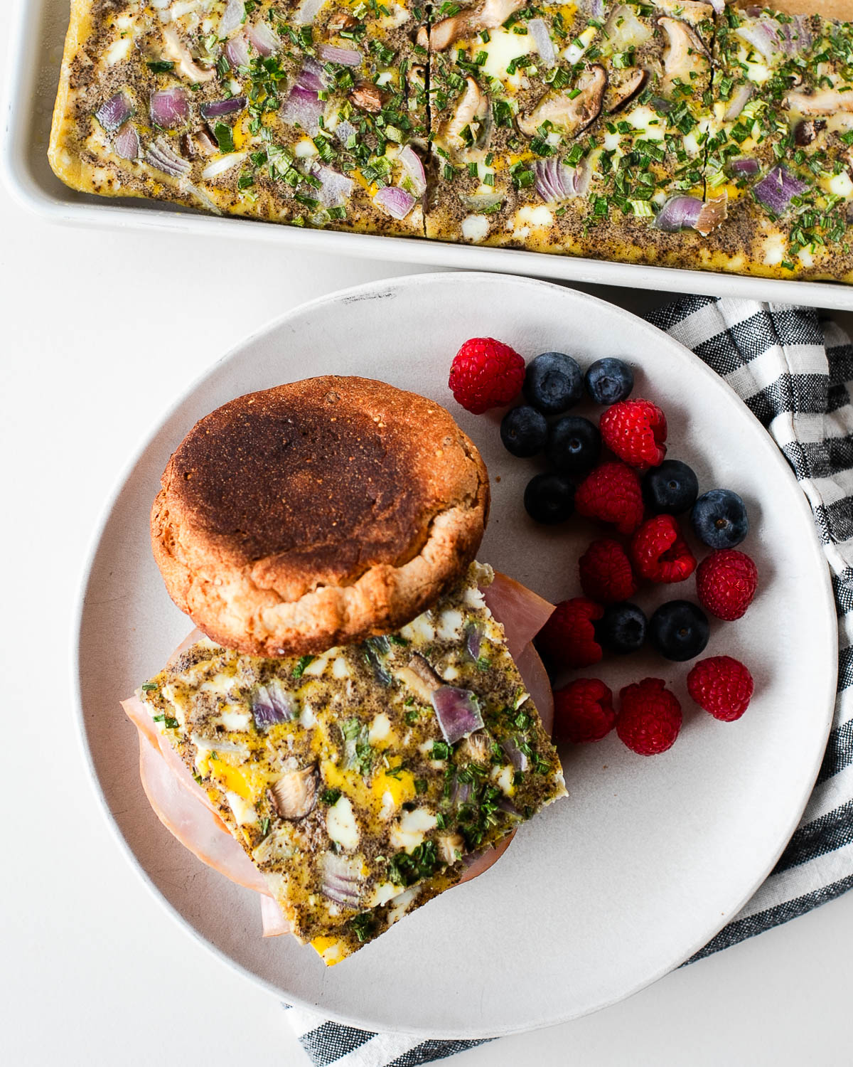 Overhead shot of a plated breakfast sandwich next to fresh berries, with the sheet pan egg bake pan visible in the background.