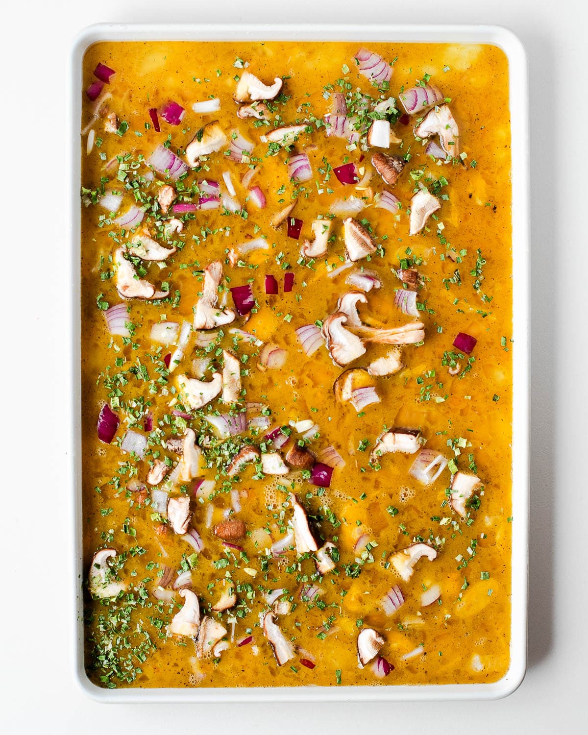 Overhead shot of raw egg mixture in a white sheet pan topped with sliced mushrooms, diced red onion, and fresh chives before baking.