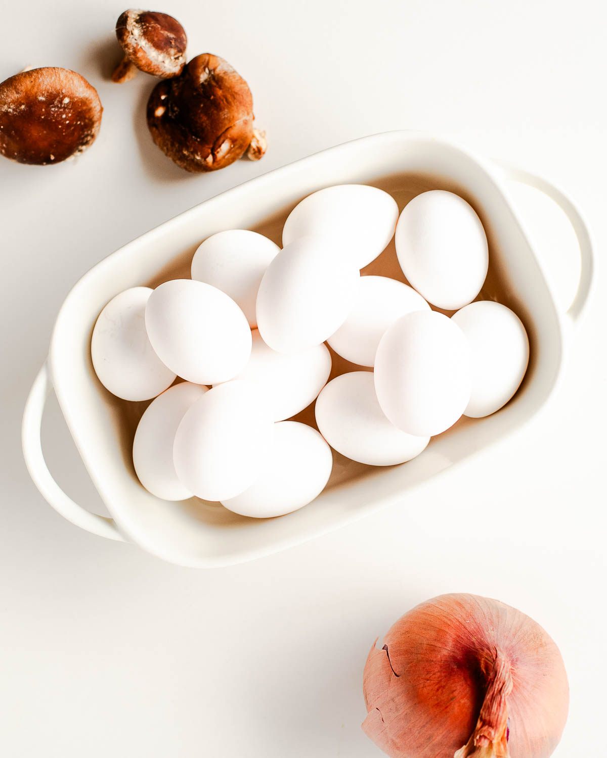 Overhead shot of whole eggs in a mixing dish alongside fresh shiitake mushrooms and a shallot, ready for the egg bake.