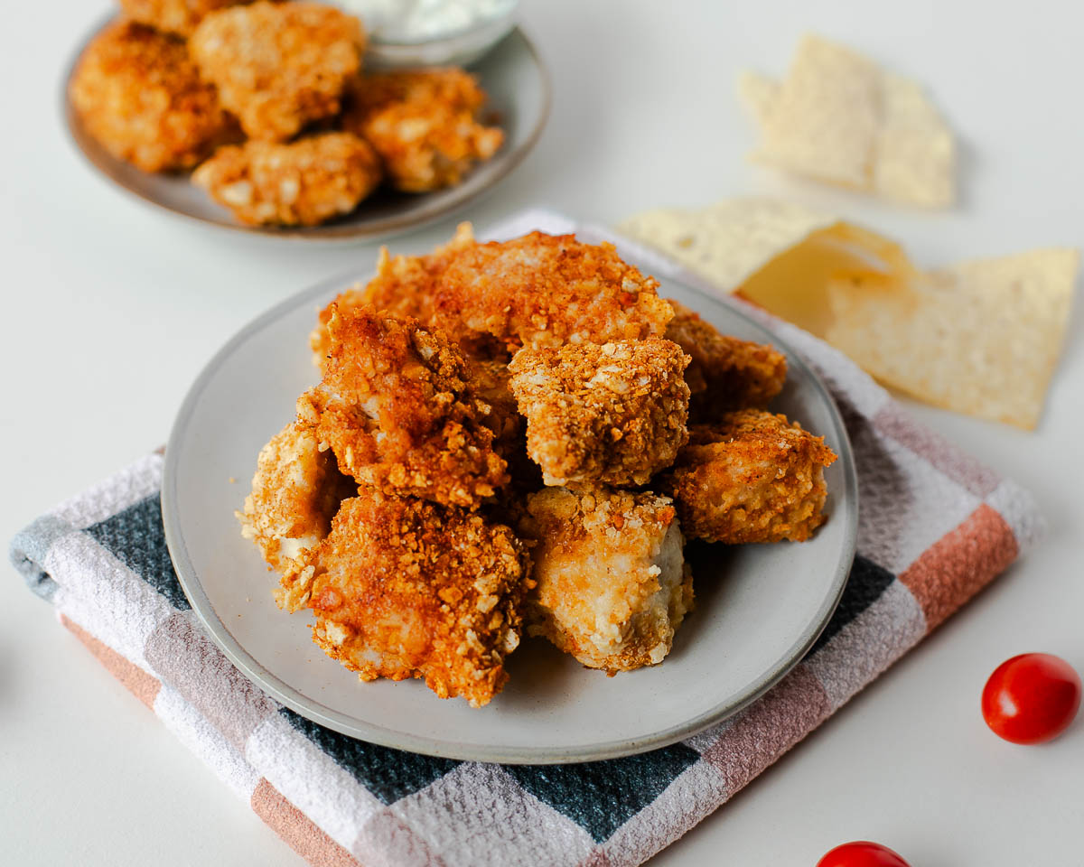 Tortilla crusted chicken nuggets on a plate with tortilla chips and cherry tomatoes in the background.