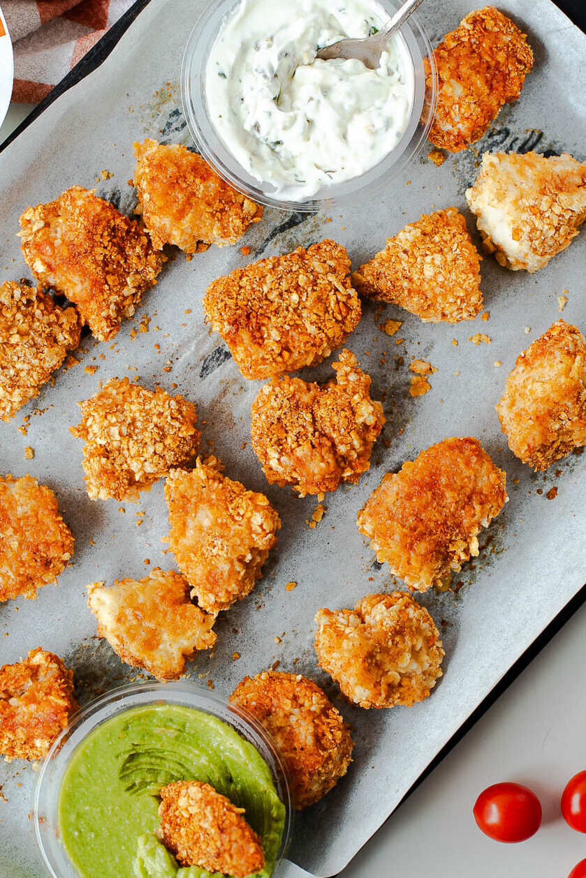 Tortilla chip crusted chicken nuggets on a baking sheet with creamy dip, guacamole, and cherry tomatoes on the side.
