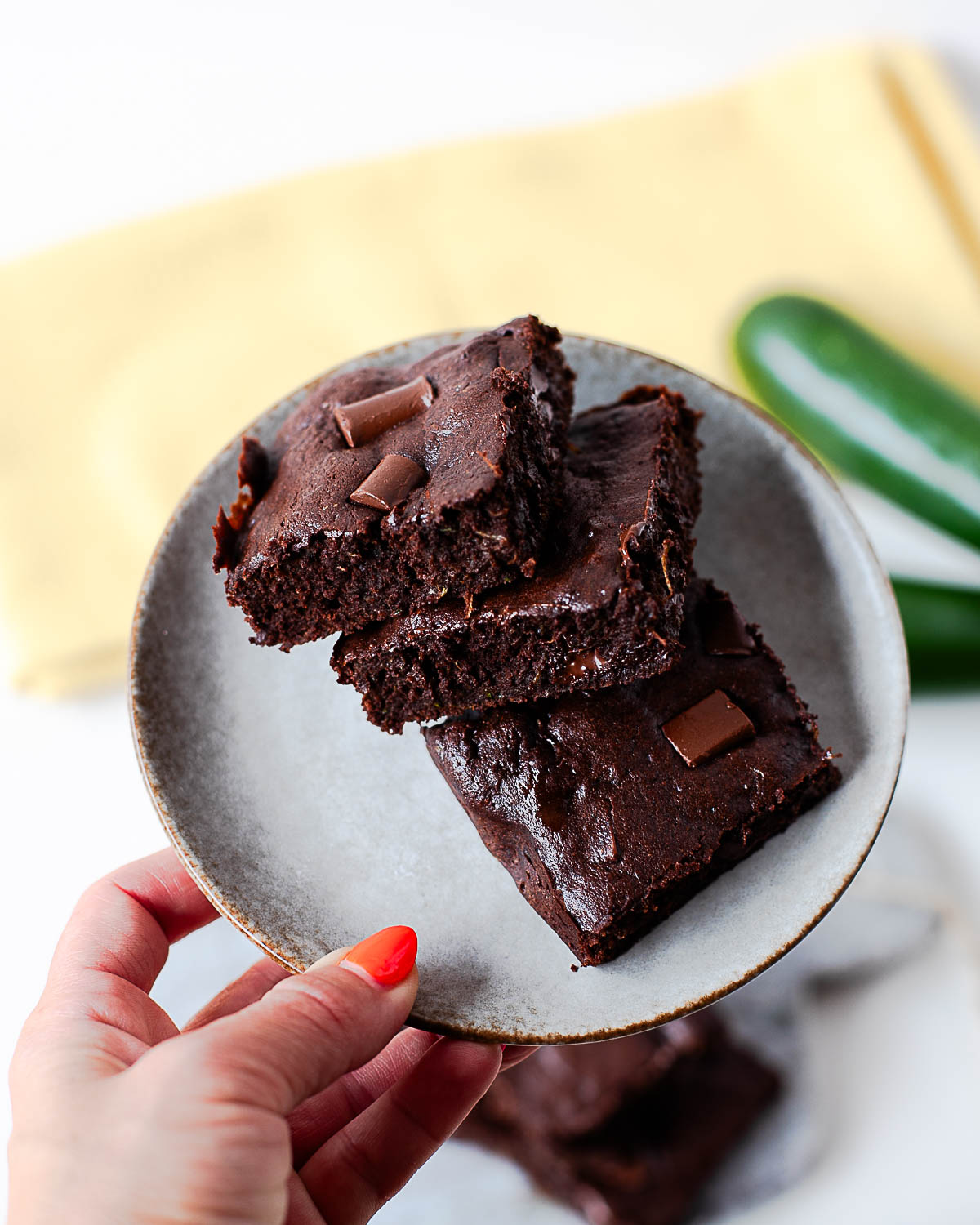 A hand holding a plate of stacked almond flour zucchini brownies, fresh zucchini in the background.