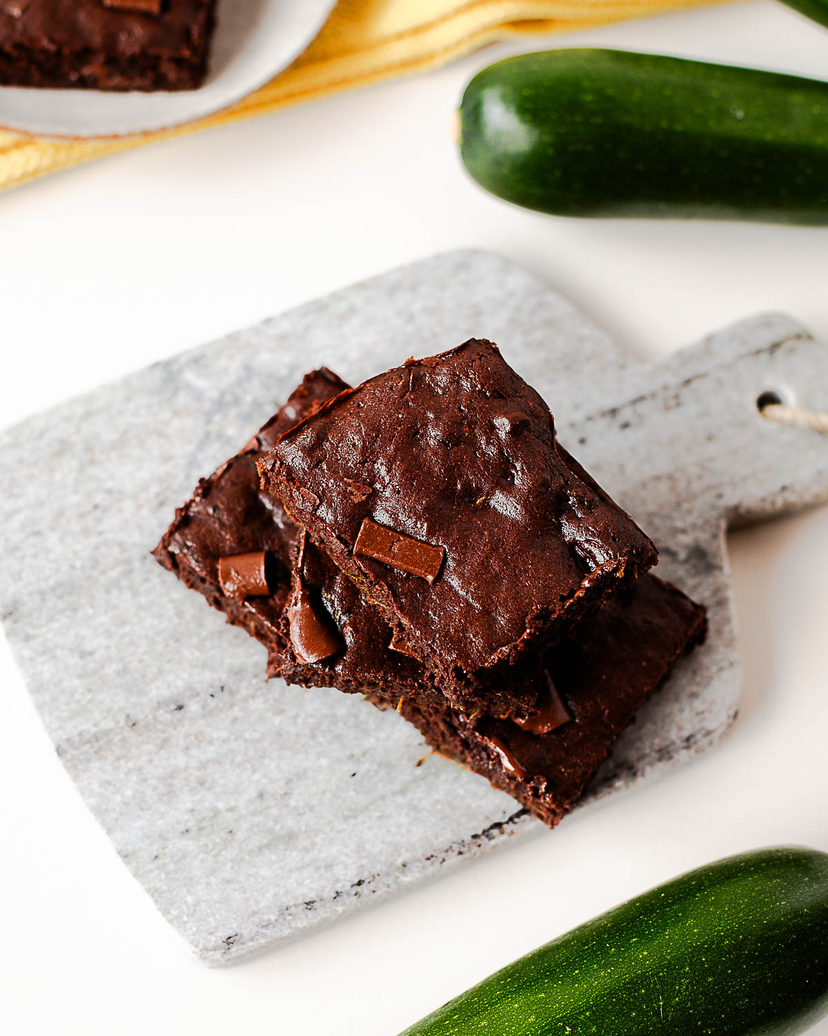 Overhead angle of stacked zucchini brownies on a marble board, with chocolate chunks and a plate of brownies nearby.