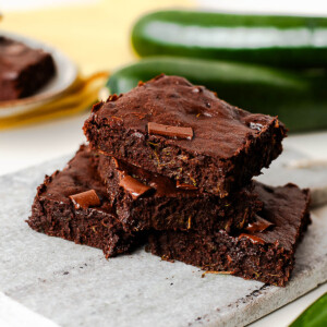 Close-up of fudgy zucchini brownies stacked on a marble board, chocolate chunks visible throughout the moist crumb.