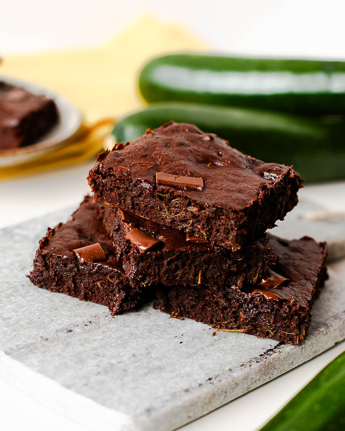 Close-up of fudgy zucchini brownies stacked on a marble board, chocolate chunks visible throughout the moist crumb.