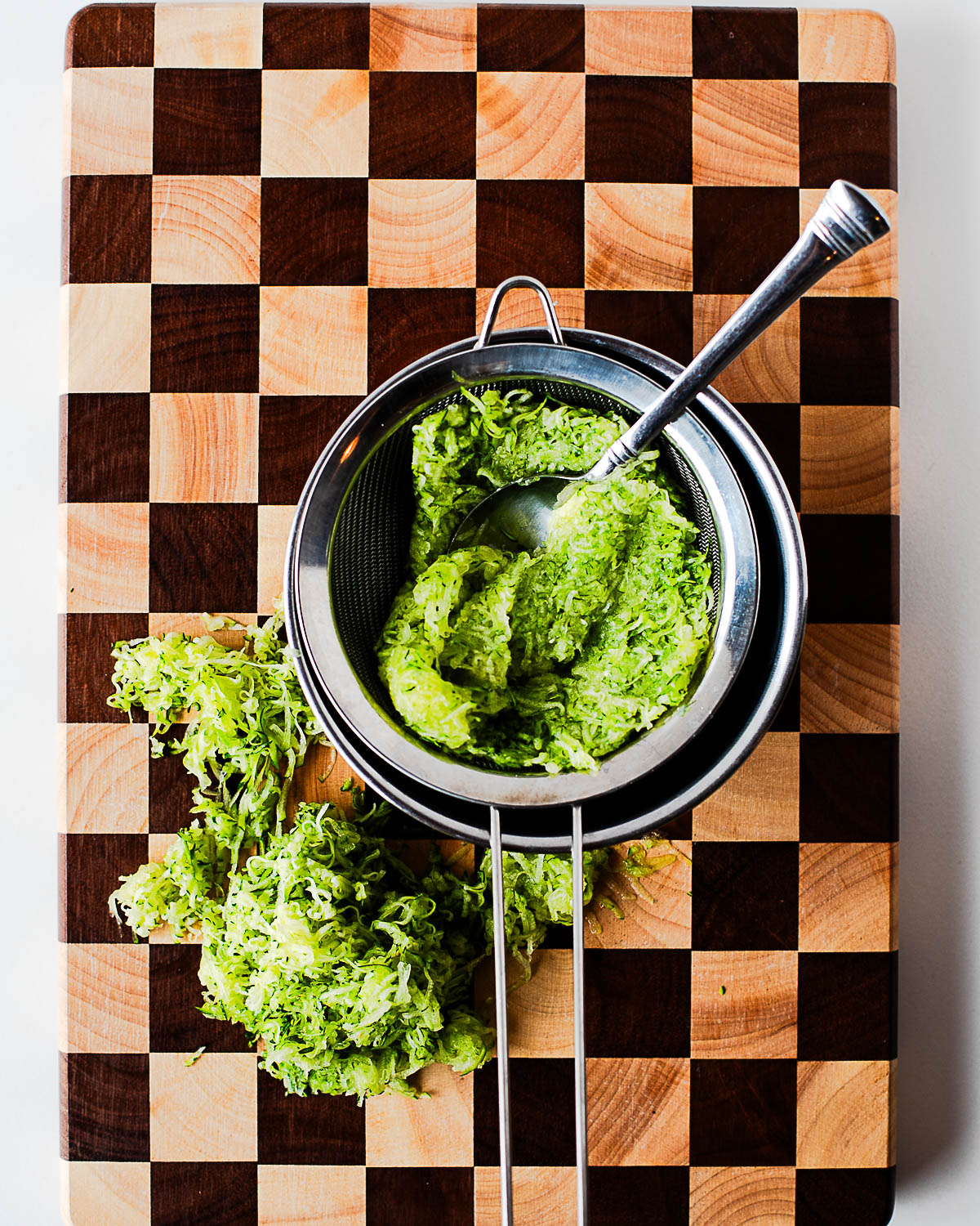 Grated zucchini being squeezed of excess liquid in a mesh strainer with a spoon, with extra shredded zucchini piled on a wooden cutting board.