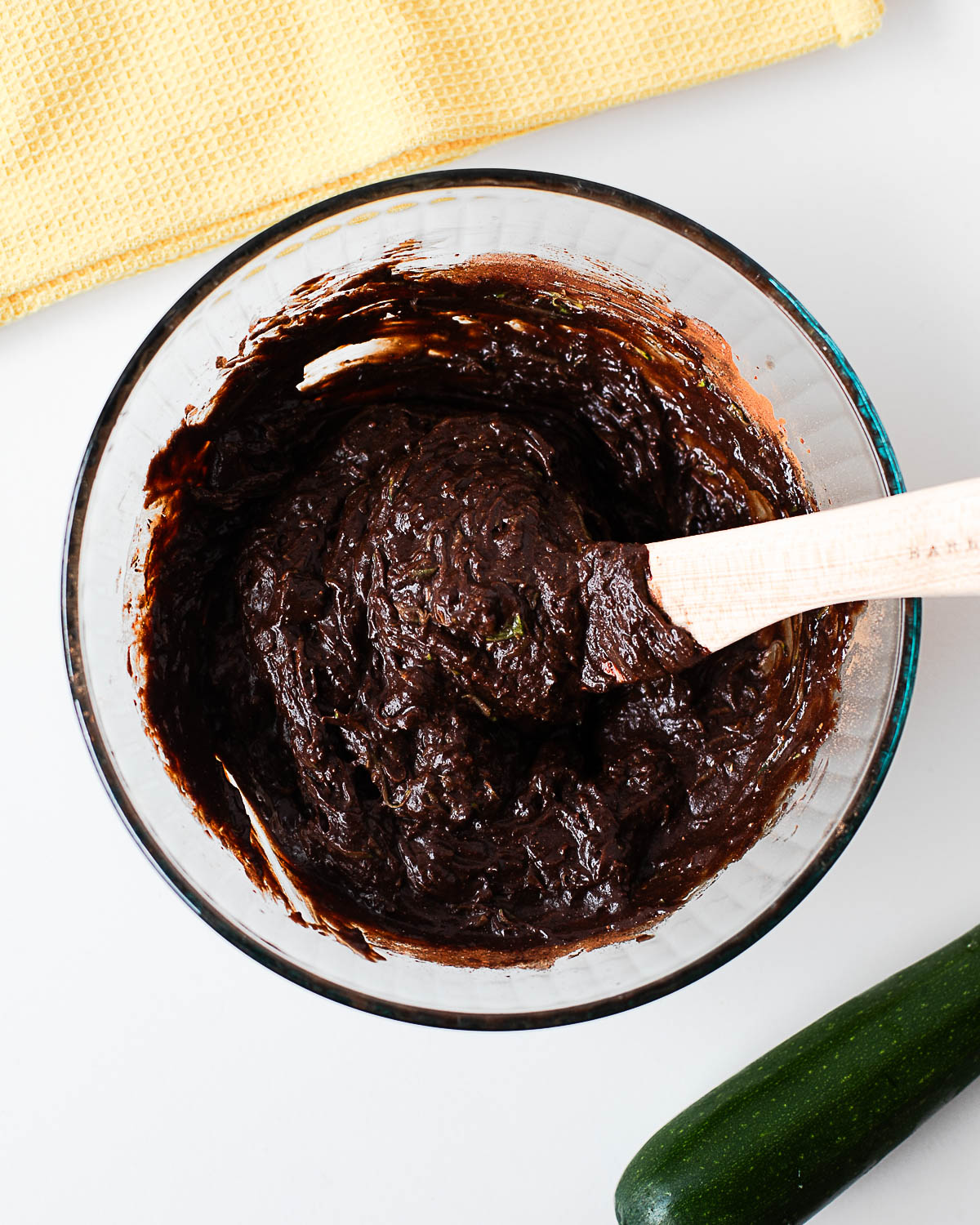 Fudgy zucchini brownie batter mixed in a glass bowl with a wooden spoon, with a whole zucchini nearby.
