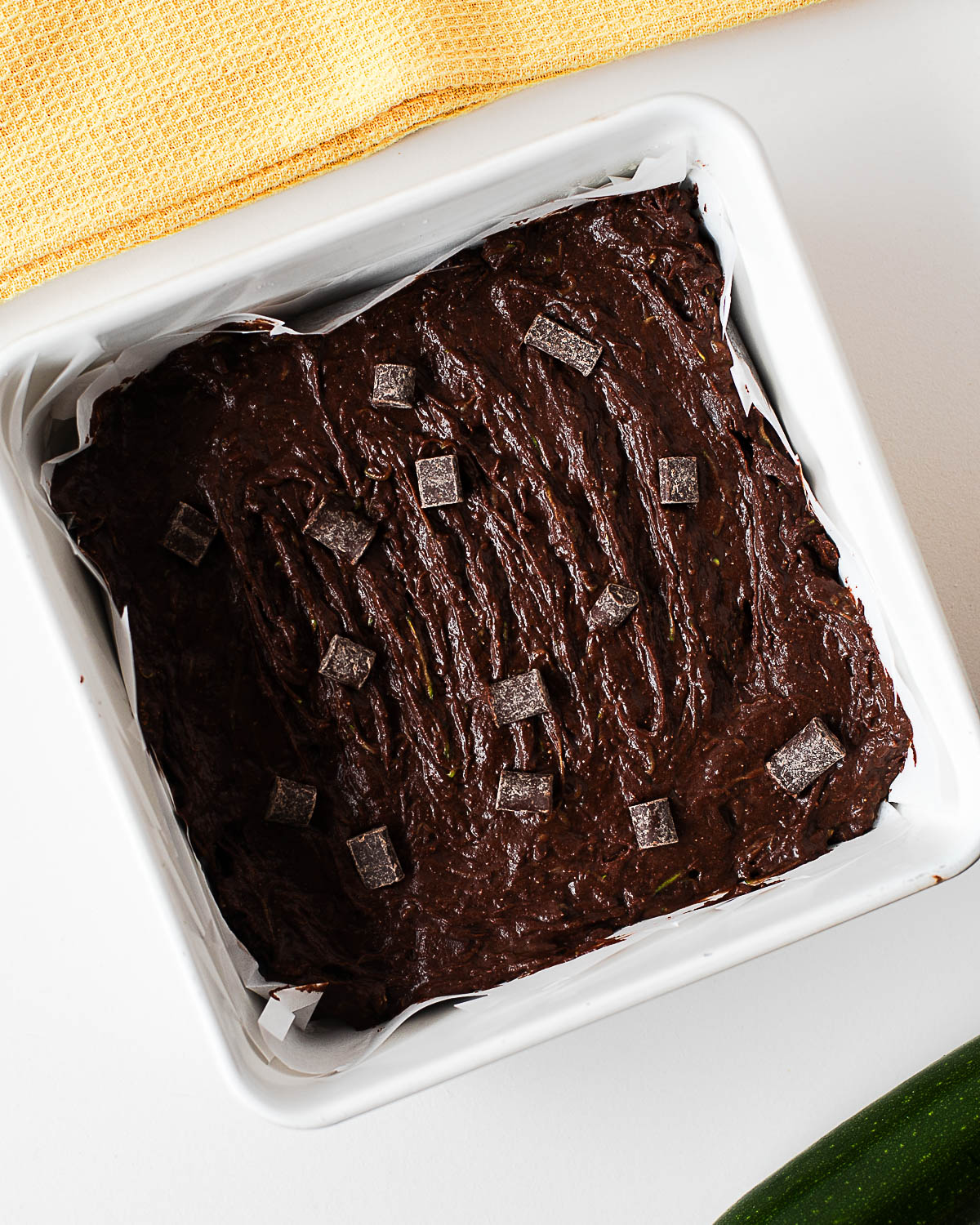 Almond flour zucchini brownie batter spread into a parchment-lined baking pan and topped with chocolate chunks before baking.