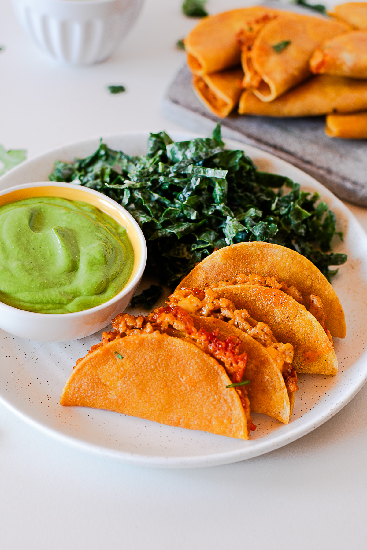 Plate of baked chicken tacos with green avocado dip and kale salad, with more tacos on a board behind