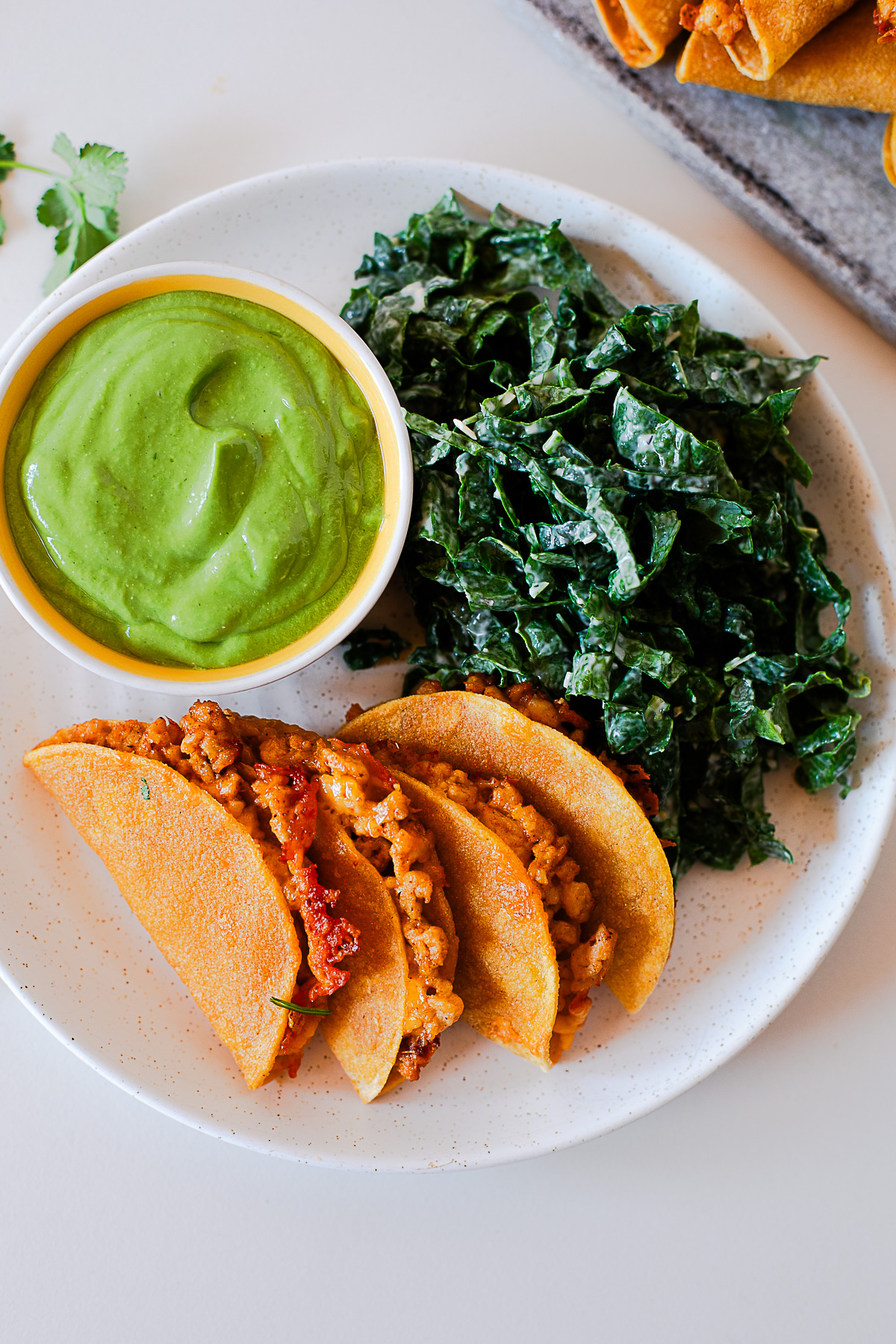 Overhead view of crispy baked chicken tacos on a plate with green avocado dip and kale salad