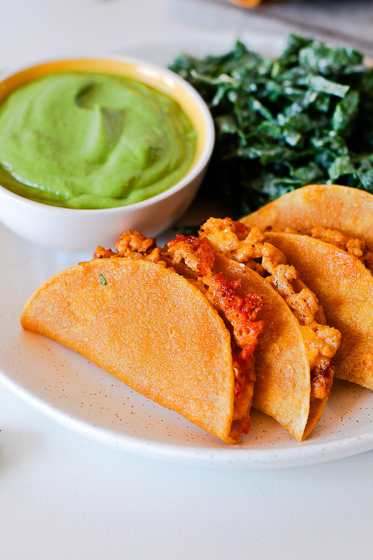 Close-up of crispy baked chicken tacos with melted cheddar next to a bowl of green avocado dip