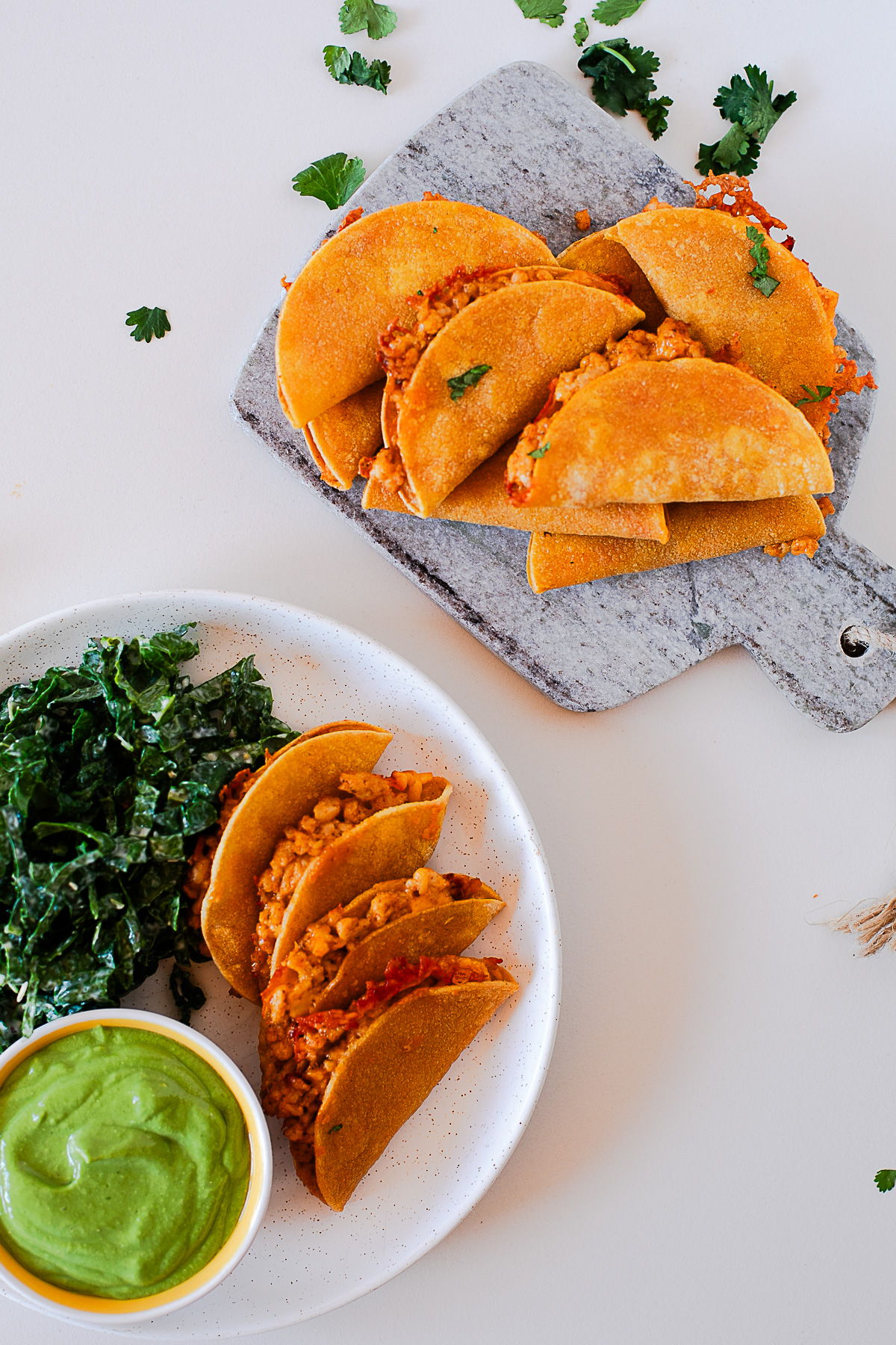 Overhead view of baked chicken tacos on a serving board and a plate with kale salad and avocado dip