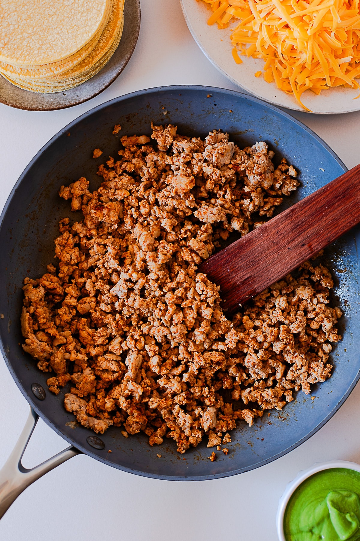 Taco seasoned ground chicken cooking in a skillet with corn tortillas and shredded cheddar nearby