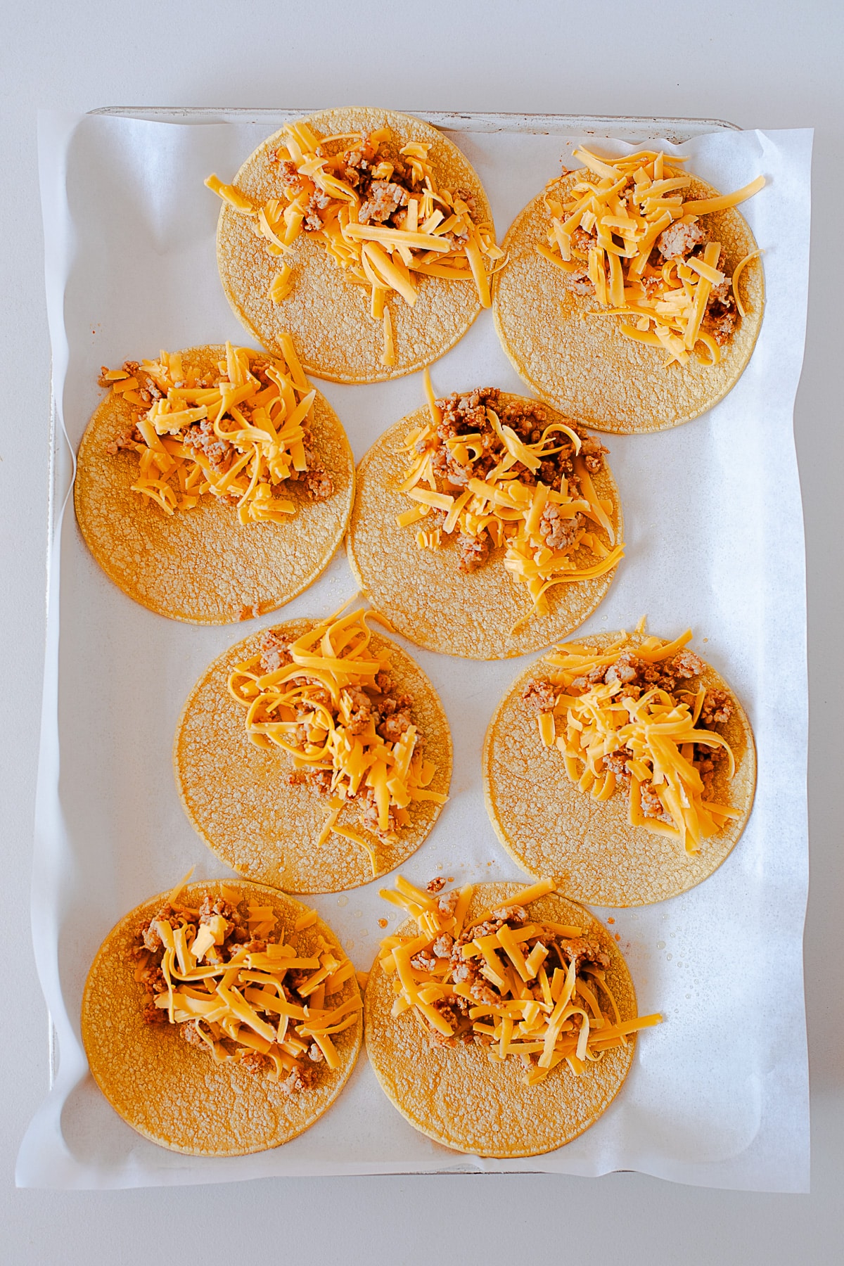 Street corn tortillas topped with shredded cheddar and seasoned ground chicken on a parchment lined baking sheet