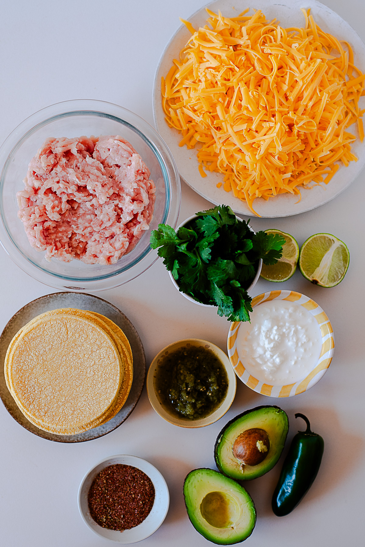Ingredients for baked chicken tacos and green avocado dip, including ground chicken, cheddar, corn tortillas, cilantro, cottage cheese, salsa verde, avocado and jalapeño