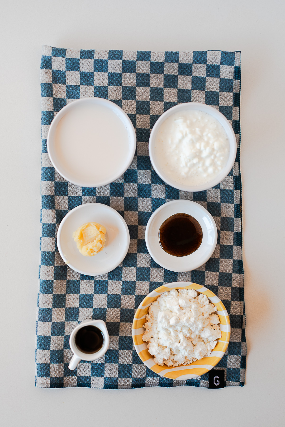 Overhead shot of the recipe ingredients, including almond milk, cottage cheese, browned butter ghee, and vanilla bean paste, arranged on a checkered towel.