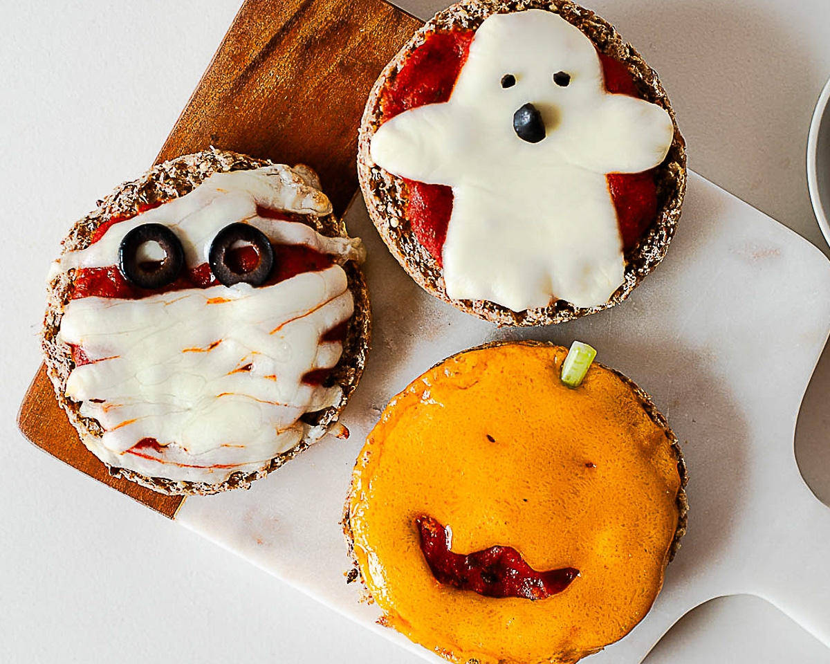 Three Halloween mini pizzas on a cutting board: a mummy pizza, a ghost pizza, and a jack-o'-lantern pizza with a green onion stem.