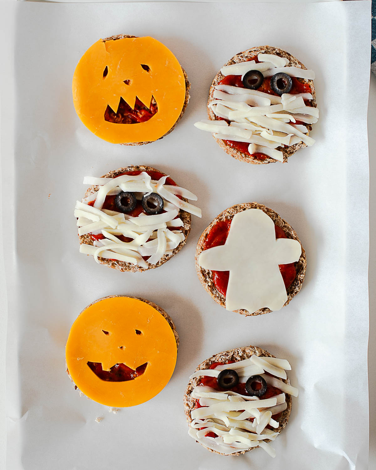 Six unbaked Halloween mini pizzas on a parchment-lined baking sheet, including jack-o'-lantern, mummy, and ghost shapes made from cheese.