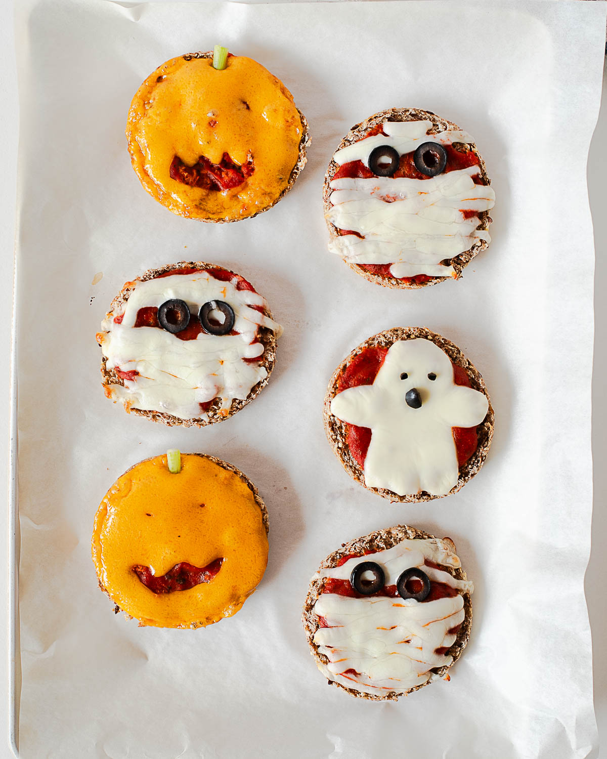 Six baked Halloween mini pizzas on a baking sheet with melted cheese, showing jack-o'-lantern, mummy, and ghost designs.
