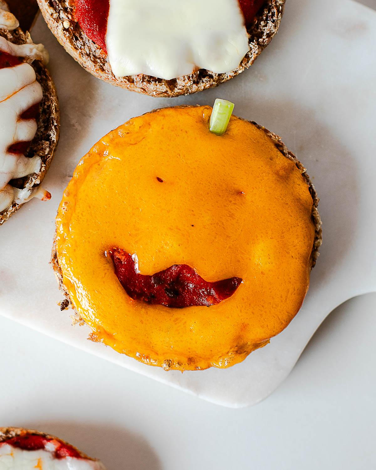 Close-up of a jack-o'-lantern Halloween mini pizza made with a cheddar cheese face and a green onion stem.