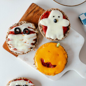 Three Halloween mini pizzas arranged on a cutting board, showing mummy, ghost, and jack-o'-lantern designs.