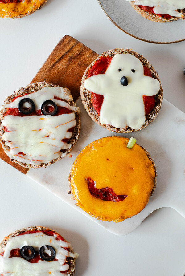 Three Halloween mini pizzas arranged on a cutting board, showing mummy, ghost, and jack-o'-lantern designs.