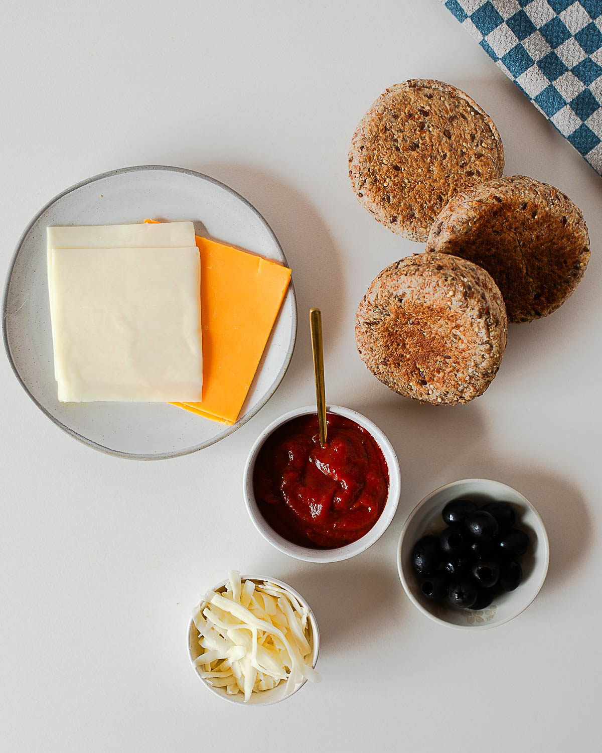 Flat lay of ingredients for Halloween mini pizzas: cheese slices, shredded mozzarella, pizza sauce, black olives, and English muffins.
