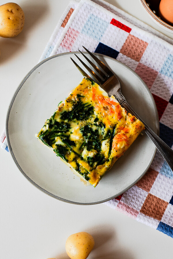 A slice of smoked salmon breakfast casserole on a gray plate with a fork, showing the potato layer underneath.